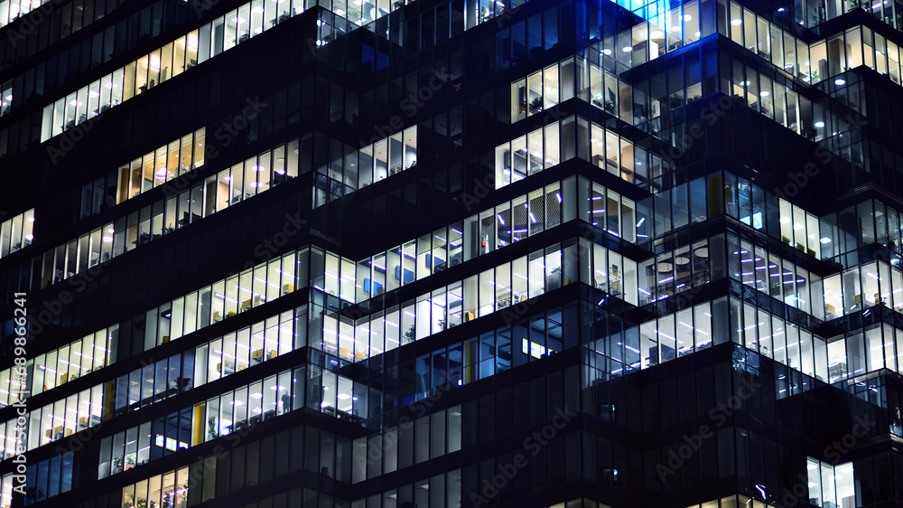 Obraz premium Fragment of the glass facade of a modern corporate building at night. Modern glass office in city. Big glowing windows in modern office buildings at night, in rows of windows light shines. 