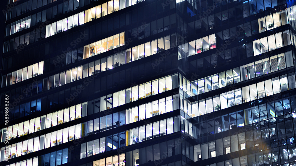 Obraz premium Fragment of the glass facade of a modern corporate building at night. Modern glass office in city. Big glowing windows in modern office buildings at night, in rows of windows light shines. 