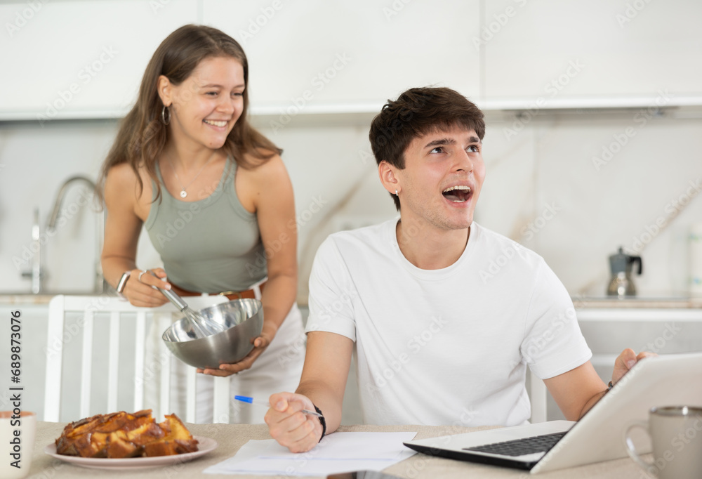 Young woman smiling and talking to young guy working at laptop in kitchen