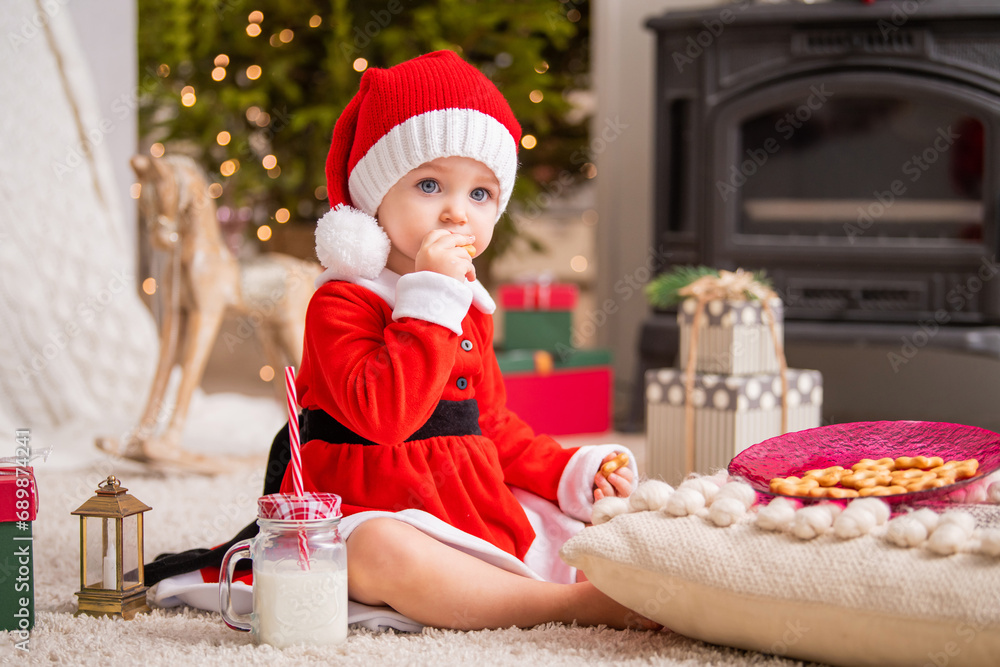 Cheerful little girl dressed as Santa Claus eats cookies and drinks milk in the living room on the background of the Christmas tree. Cheerful little girl dressed as Santa Claus eats cookies and drinks milk in the living room on the background of the Christmas tree.