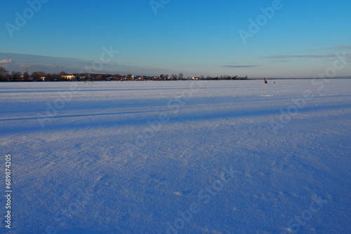 Frozen Lake Logmozero, Karelia, Russia. Winter January in nature. Smooth blue ice surface covered with snow. Evening winter sunset in sunny weather. Horizon with coast. Blue sky