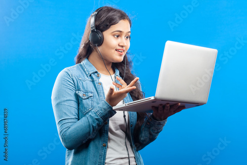 Caucasian female with headphones holding laptop and busy in a video call, isolated on blue background
