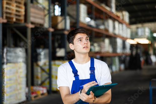 Young man conducts verification, examination and inspection of goods in warehouse and records work data in paper report
