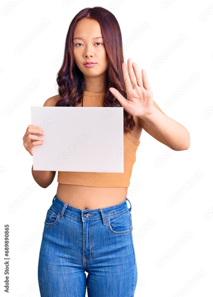 Young beautiful chinese girl holding paper banner with blank space with open hand doing stop sign with serious and confident expression, defense gesture Young beautiful chinese girl holding paper banner with blank space with open hand doing stop sign with serious and confident expression, defense gesture