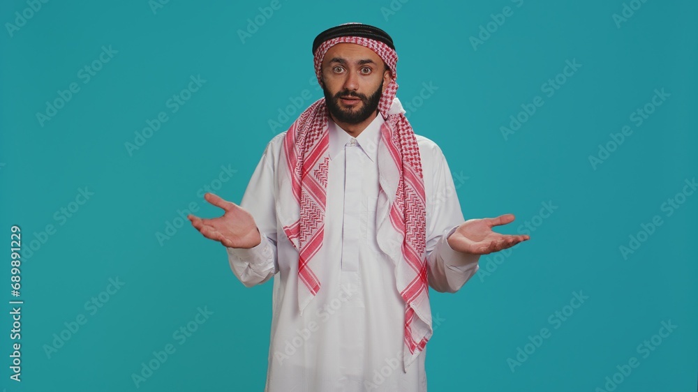 Islamic adult acting unsure and clueless in studio, showcasing i dont know sign and being confused over blue background. Young person in arab clothing feeling uncertain and shrugging. Islamic adult acting unsure and clueless in studio, showcasing i dont know sign and being confused over blue background. Young person in arab clothing feeling uncertain and shrugging.