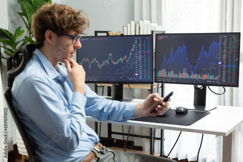 Young investor stock trader looking on smartphone, analyzing dynamic financial exchange investment on two laptop screens background in current market graph in real time at modern office. Gusher.