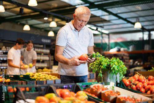 Elderly man shopper choosing cucumbers and tomatoes in grocery store