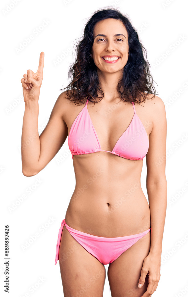 Young beautiful hispanic woman wearing bikini showing and pointing up with finger number one while smiling confident and happy. Young beautiful hispanic woman wearing bikini showing and pointing up with finger number one while smiling confident and happy.