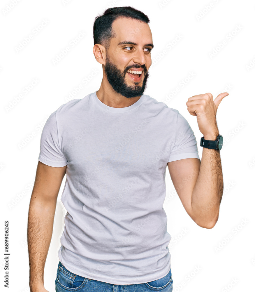 Young man with beard wearing casual white t shirt smiling with happy face looking and pointing to the side with thumb up. Young man with beard wearing casual white t shirt smiling with happy face looking and pointing to the side with thumb up.