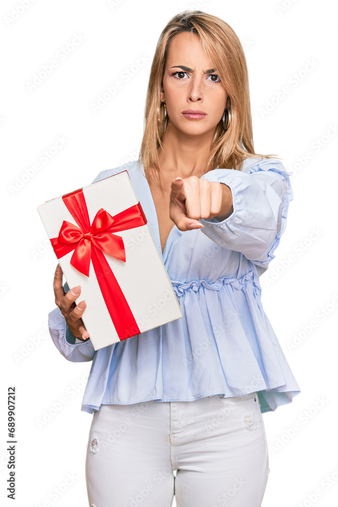 Beautiful blonde woman holding gift pointing with finger to the camera and to you, confident gesture looking serious Beautiful blonde woman holding gift pointing with finger to the camera and to you, confident gesture looking serious