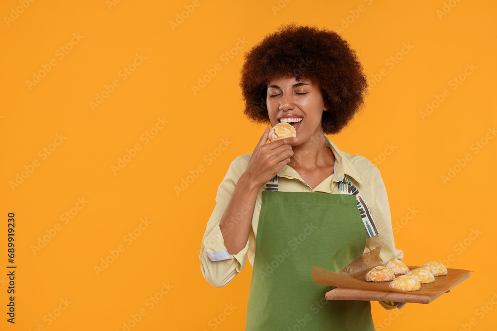 Happy young woman in apron holding board with cookies on orange background. Space for text Happy young woman in apron holding board with cookies on orange background. Space for text