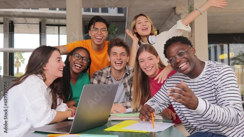 Group of young teenage students waving hands at library, using a laptop, looking at camera. Happy classmates studying and doing homeworks at highschool. Multiracial academy people staring front. High photo