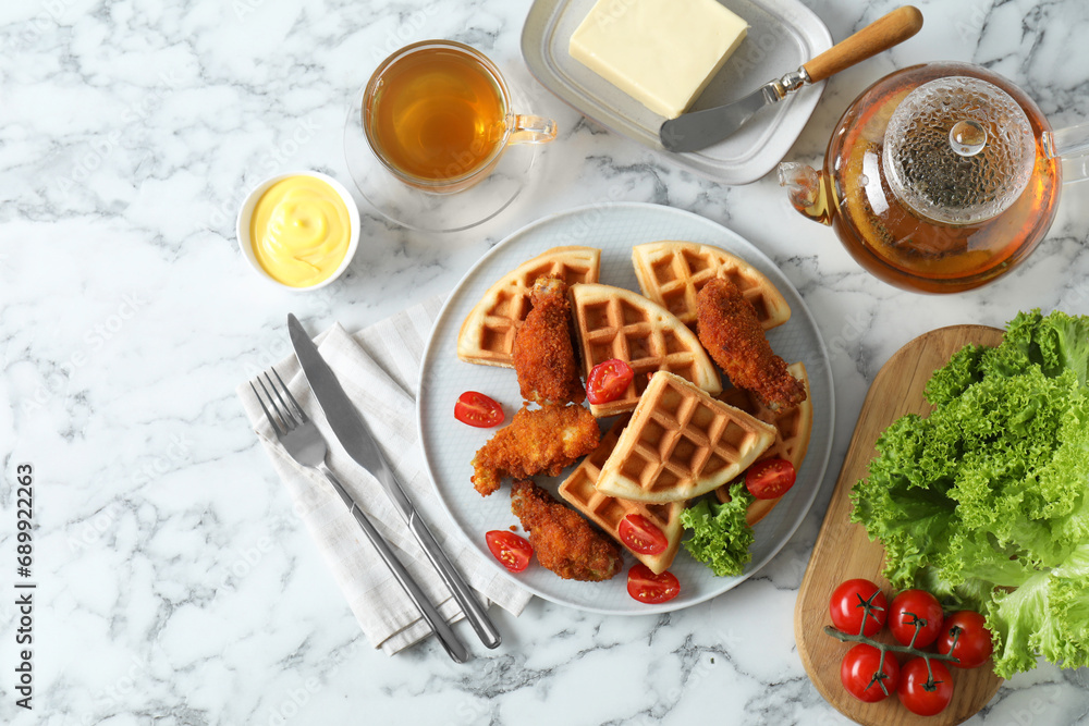Tasty Belgian waffles served with fried chicken, tomatoes, lettuce and tea on white marble table, flat lay