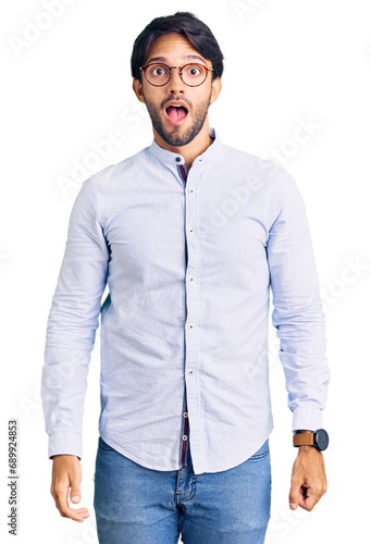 Handsome hispanic man wearing business shirt and glasses in shock face, looking skeptical and sarcastic, surprised with open mouth