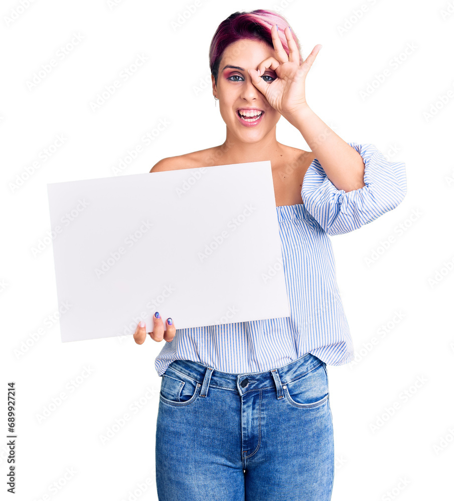 Young beautiful woman with pink hair holding cardboard banner with blank space smiling happy doing ok sign with hand on eye looking through fingers Young beautiful woman with pink hair holding cardboard banner with blank space smiling happy doing ok sign with hand on eye looking through fingers