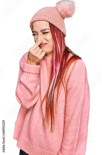 Young caucasian woman wearing casual clothes and wool cap smelling something stinky and disgusting, intolerable smell, holding breath with fingers on nose. bad smell