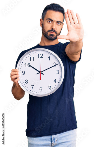 Young hispanic man holding big clock with open hand doing stop sign with serious and confident expression, defense gesture