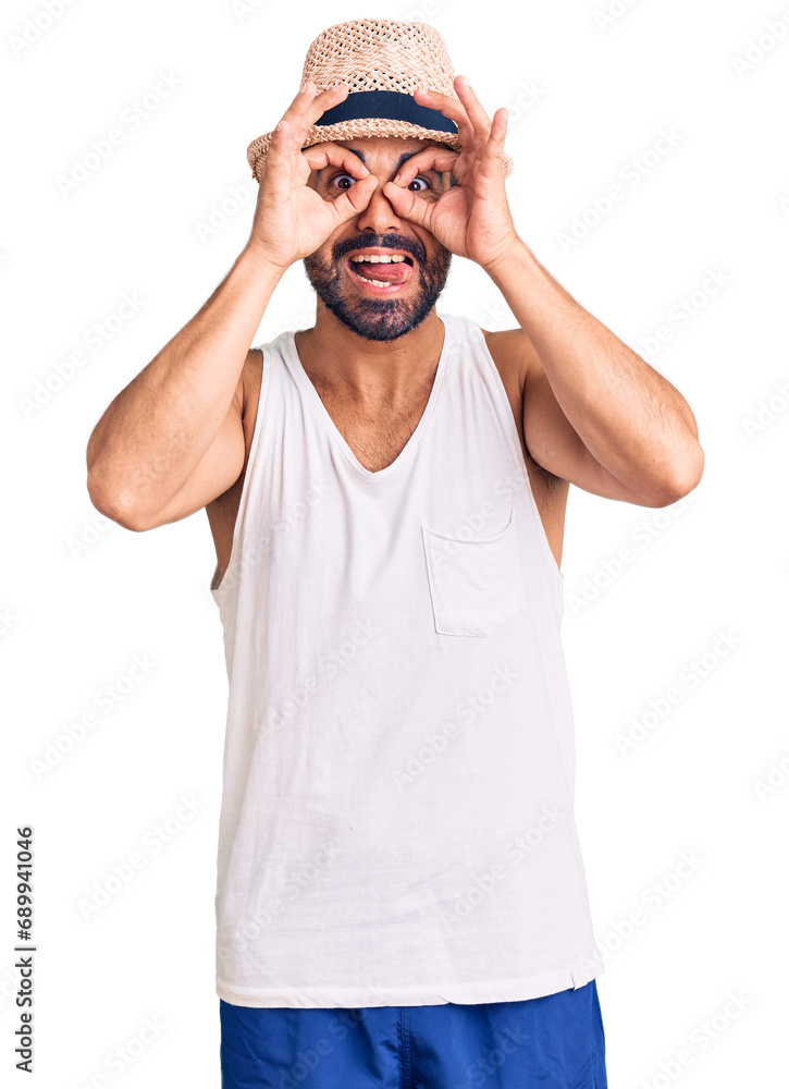 Fototapeta premium Young hispanic man wearing casual summer hat doing ok gesture like binoculars sticking tongue out, eyes looking through fingers. crazy expression.