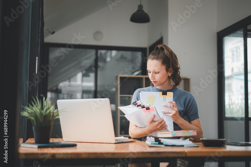 Young busy business woman manager, lawyer or company employee holding accounting bookkeeping documents checking financial data or marketing report working in office with laptop. Paperwork