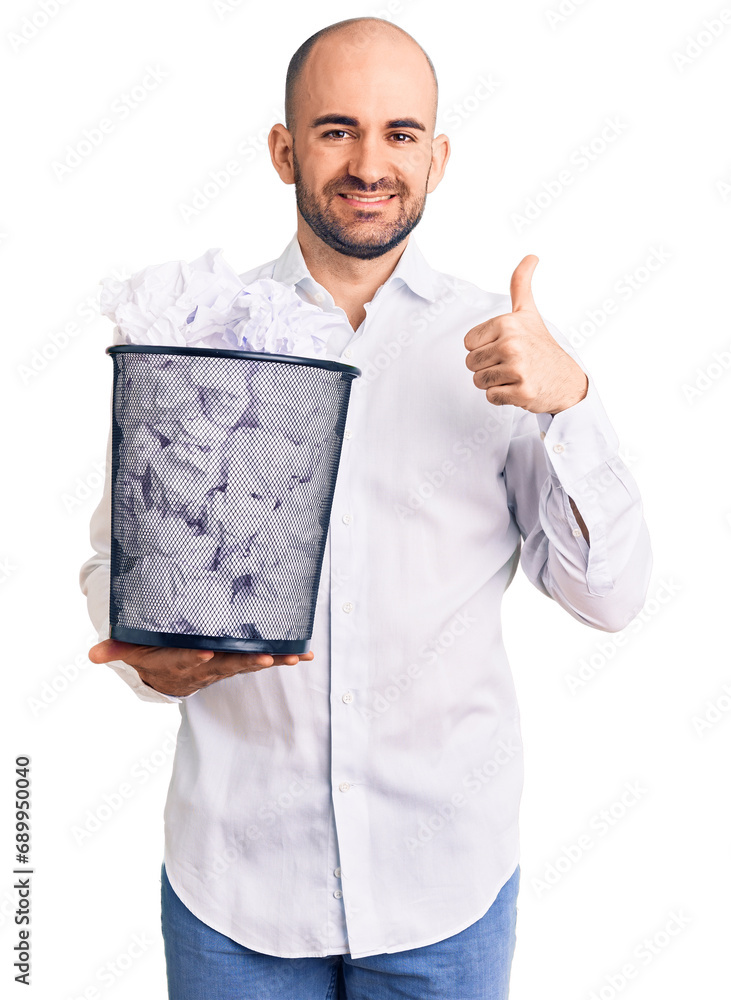 Young handsome man holding paper bin full of crumpled papers smiling happy and positive, thumb up doing excellent and approval sign Young handsome man holding paper bin full of crumpled papers smiling happy and positive, thumb up doing excellent and approval sign