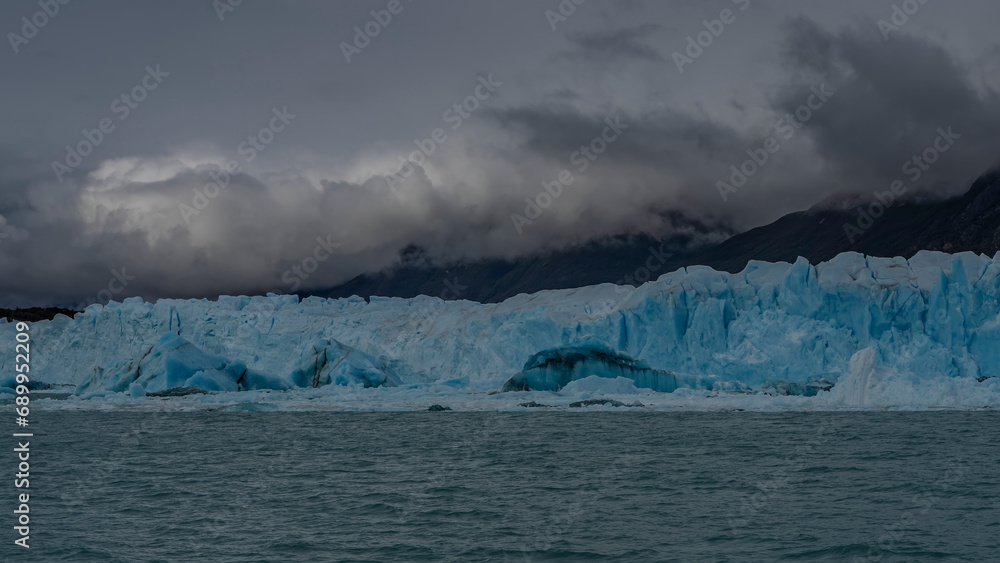 Perito Moreno Glacier. A wall of blue ice with cracks, sharp peaks against a cloudy sky and mountains. Melting ice floes float in the turquoise water of the lake. Argentina. El Calafate.Lago Argentino