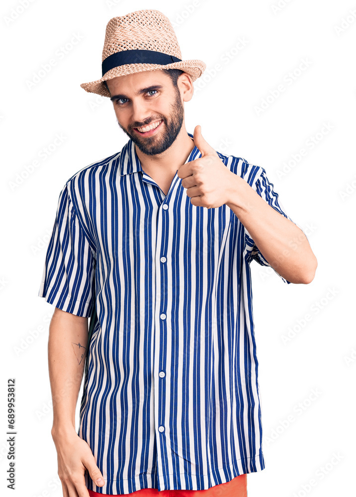 Young handsome man with beard wearing summer hat and striped shirt doing happy thumbs up gesture with hand. approving expression looking at the camera showing success. Young handsome man with beard wearing summer hat and striped shirt doing happy thumbs up gesture with hand. approving expression looking at the camera showing success.