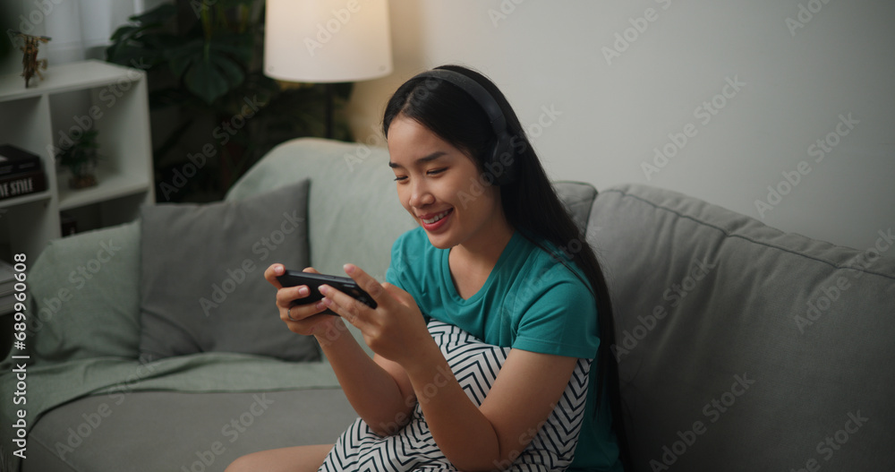 Portrait of Young Asian woman wearing glasses and headphones enjoys playing online esport games on smartphone sitting on sofa in the living room at home,Gamer lifestyle concept. Portrait of Young Asian woman wearing glasses and headphones enjoys playing online esport games on smartphone sitting on sofa in the living room at home,Gamer lifestyle concept.