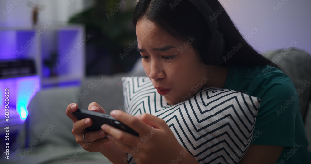 Portrait of Young Asian woman wearing glasses and headphones enjoys playing online esport games on smartphone sitting on sofa in the living room at home,Gamer lifestyle concept.