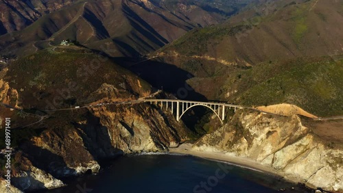 4 k drone footage San Francisco, California. Aerial Shot Of Bixby Canyon Bridge At Beach, Drone Flying Backwards Over the ocean offering views of green mountains at evening sun.