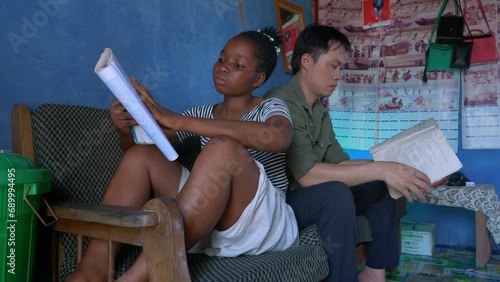 back to back sitting couple mixed race while reading a book on a couch inside a house in remote village