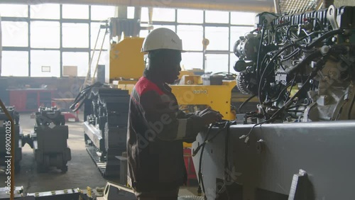Side panning shot of Black factory worker in overalls standing and performing diagnosis of caterpillar tractor internal explosion engine at machinery plant photo