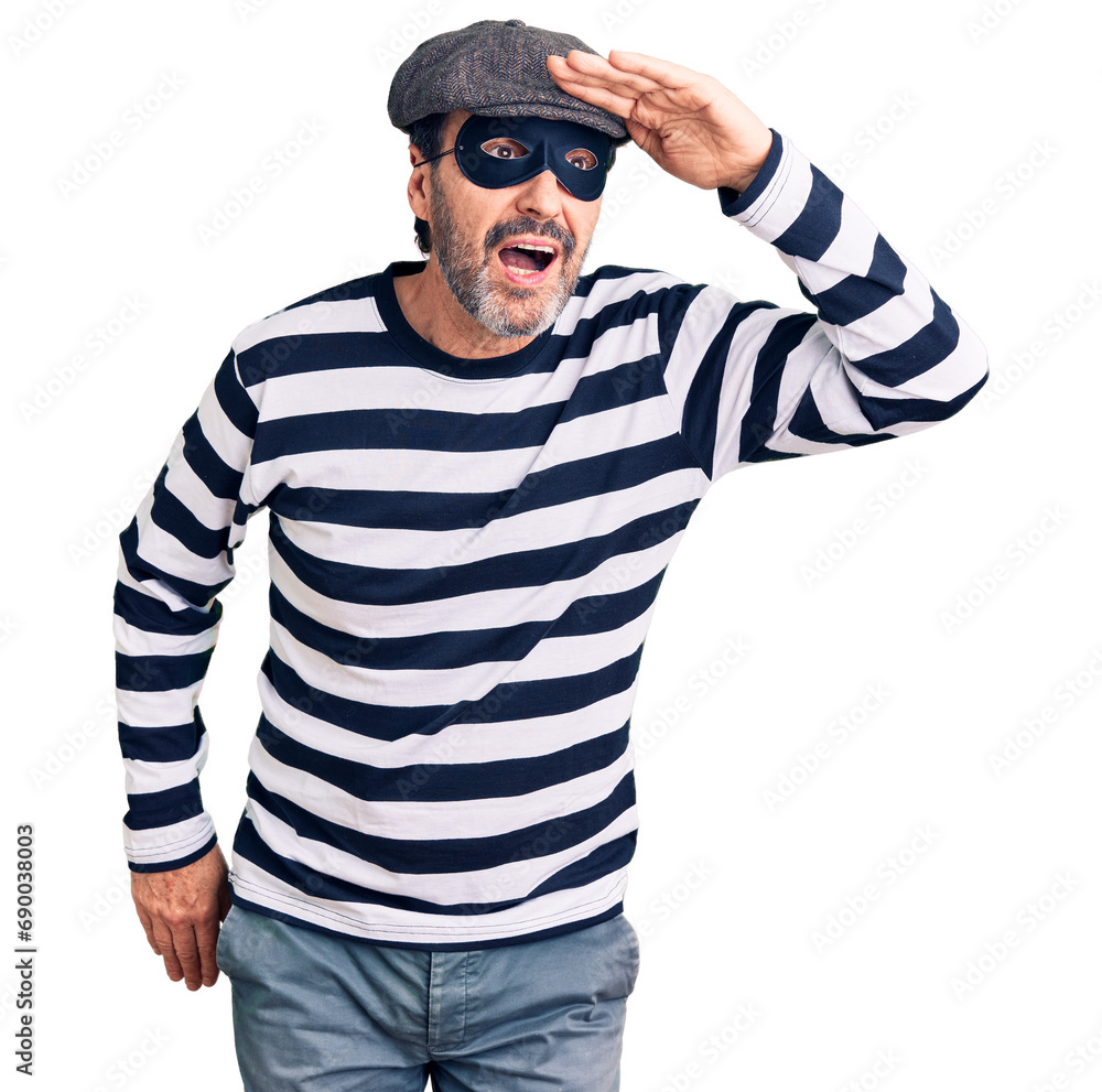 Middle age handsome man wearing burglar mask very happy and smiling looking far away with hand over head. searching concept. Middle age handsome man wearing burglar mask very happy and smiling looking far away with hand over head. searching concept.