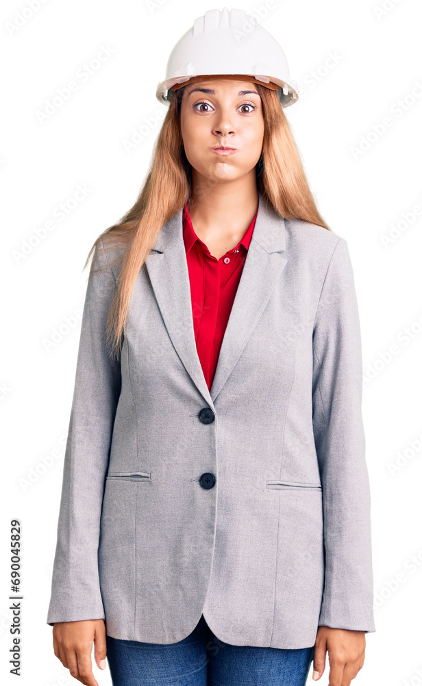 Beautiful young woman wearing architect hardhat puffing cheeks with funny face. mouth inflated with air, crazy expression. Beautiful young woman wearing architect hardhat puffing cheeks with funny face. mouth inflated with air, crazy expression.