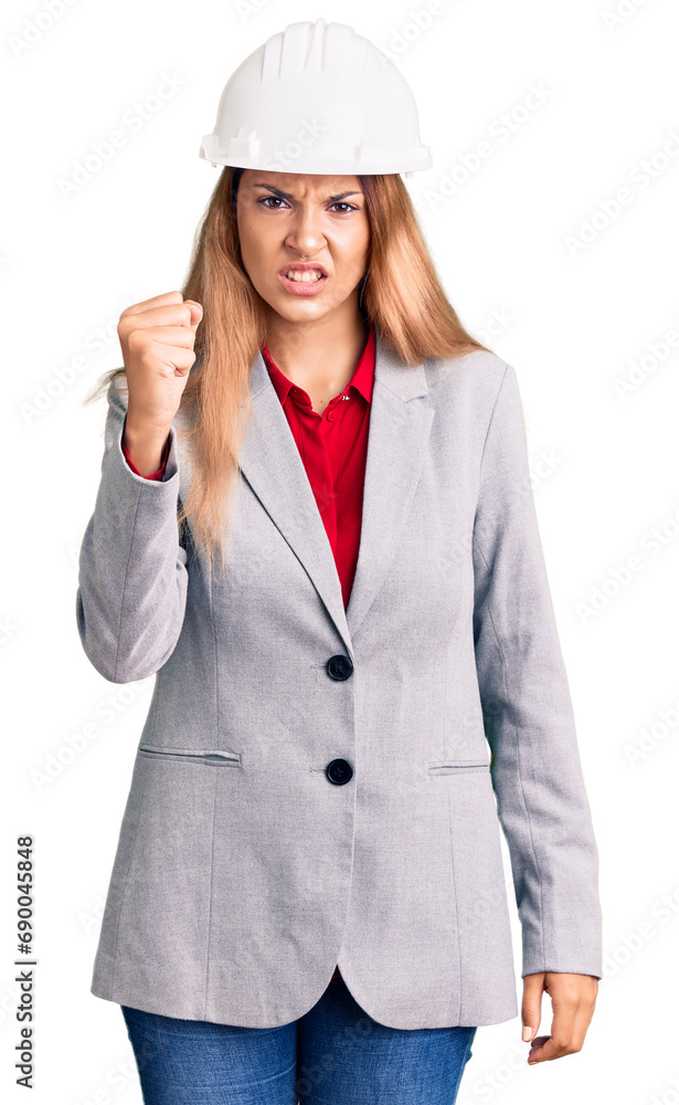 Beautiful young woman wearing architect hardhat angry and mad raising fist frustrated and furious while shouting with anger. rage and aggressive concept. Beautiful young woman wearing architect hardhat angry and mad raising fist frustrated and furious while shouting with anger. rage and aggressive concept.
