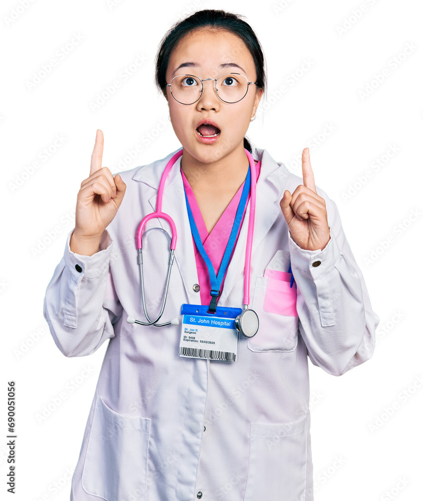 Young chinese woman wearing doctor uniform and stethoscope amazed and surprised looking up and pointing with fingers and raised arms. Young chinese woman wearing doctor uniform and stethoscope amazed and surprised looking up and pointing with fingers and raised arms.