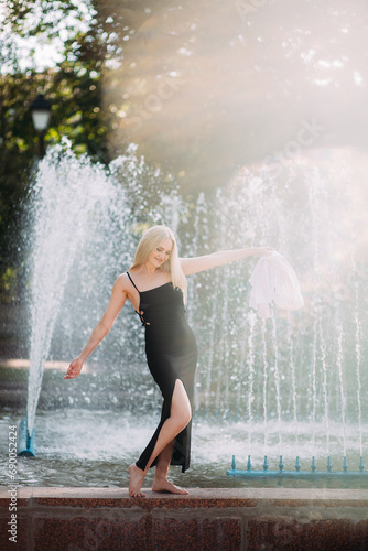 A beautiful smiling slender woman with long blond hair, in a black long dress and a white shirt, walks along the fountain in the city park, on a bright sunny day.