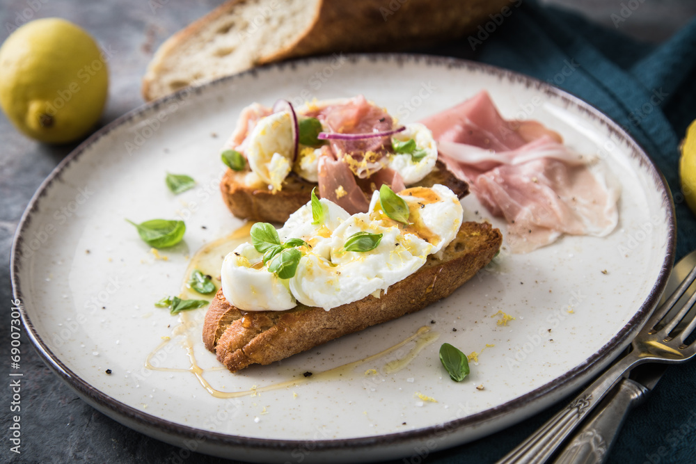 Italienische Burrata Käse Crostini aus Ciabatta Brot,konfierter Knoblauch, Prosciutto Schinken, rote Zwiebel, Basilikum, Honig, Olivenöl, Zitronen mit Teller, Besteck, Serviette blau, dunkel grau Stei