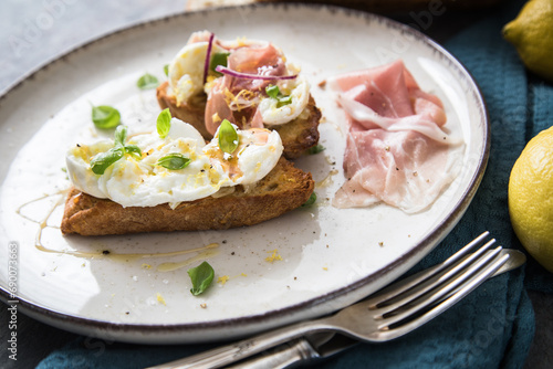 Italienische Burrata Käse Crostini aus Ciabatta Brot,konfierter Knoblauch, Prosciutto Schinken, rote Zwiebel, Basilikum, Honig, Olivenöl, Zitronen mit Teller, Besteck, Serviette blau, dunkel grau Stei