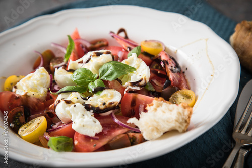 Italienischer Burrata Käse Salat mit bunten roten und gelben Tomaten, Basilikum, Ciabatta Brot Olivenöl und Balsamico Dressing mit tiefer Teller, Besteck Serviette blau, dunkel grau Stein Hintergrund