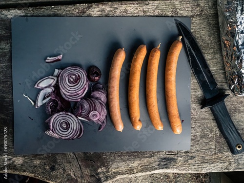 Cutting board on an outdoor wooden camping table with cut red onion, mushroom, garlic, black hunting knife, tin foil, sausages hot-dogs