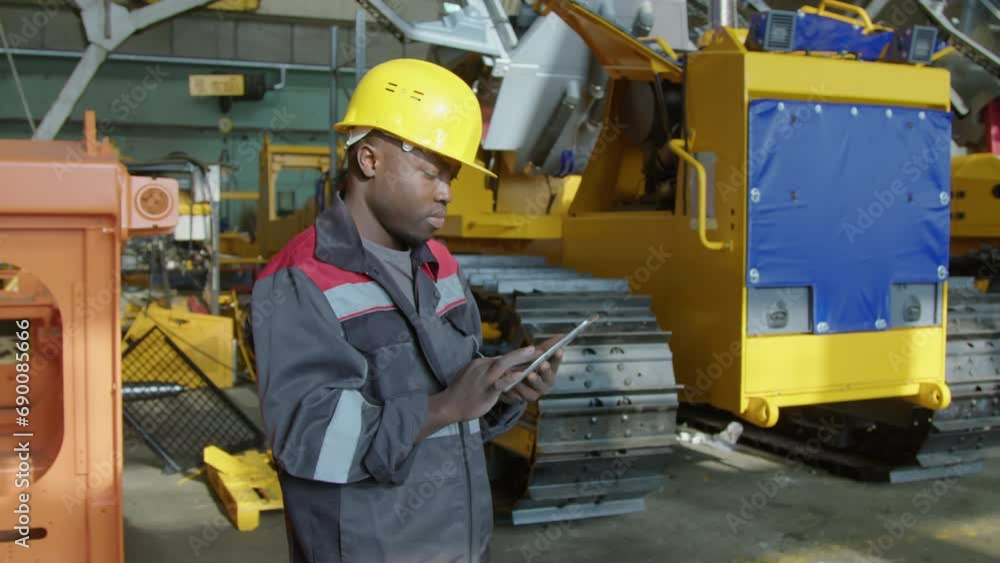 Medium arc shot of African American engineer in yellow hardhat and uniform analyzing caterpillar tractor production and typing data into tablet during workday at manufacturing plant Medium arc shot of African American engineer in yellow hardhat and uniform analyzing caterpillar tractor production and typing data into tablet during workday at manufacturing plant