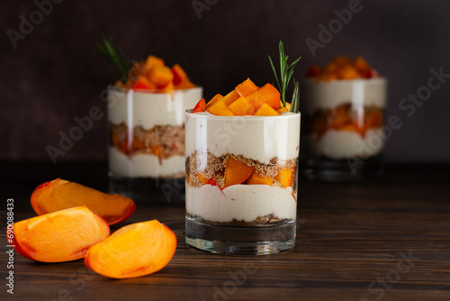 Layered dessert in a glasses with persimmon, rosemary, pecan, whipped cream and biscuit on a wooden table. Healthy food, vegan, sugar, gluten and lactose free.