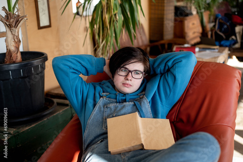 Photos Pensive androgynous teenager is lying comfortably in an armchair with a book