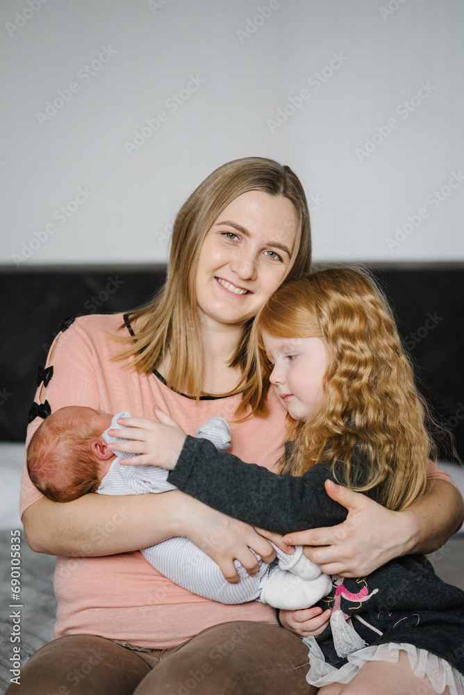 Tired mother on maternity leave.  Young mother laughing holds newborn baby son and hugs daughter. Happy mom with two children enjoying together sitting on a bed at home. Mother's day concept. Closeup.