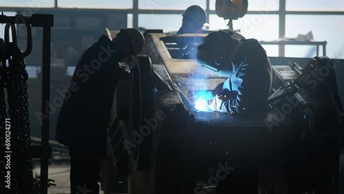 Side shot of three unidentified factory workers in welding helmets checking and welding tractor framework at machinery plant photo