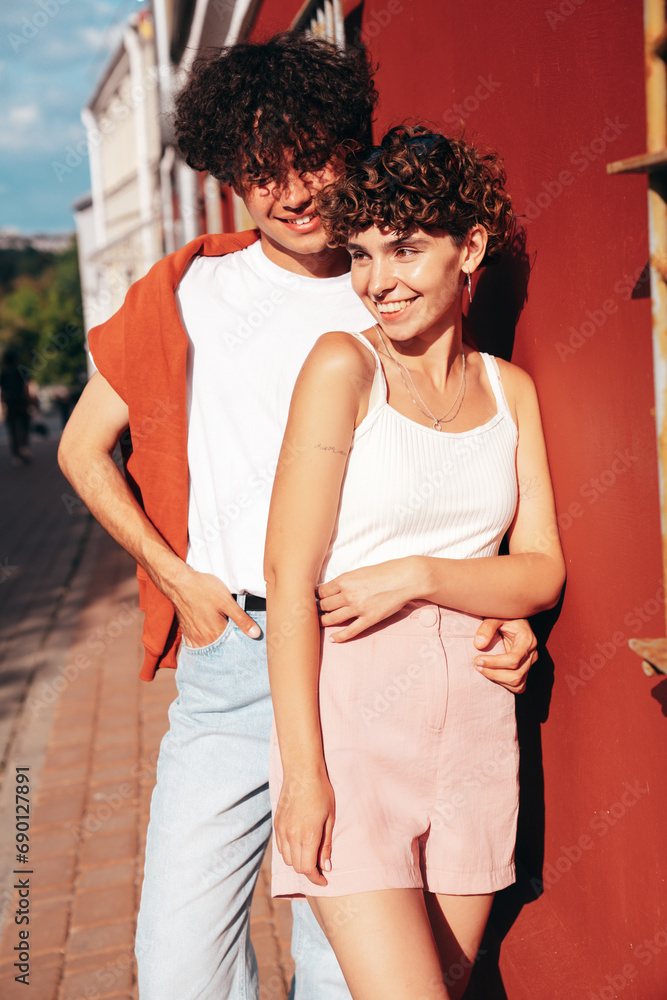 Naklejka premium Young smiling beautiful woman and her handsome boyfriend in casual summer clothes. Happy cheerful family. Female having fun. Couple posing in street at sunny day. Near wall at sunset