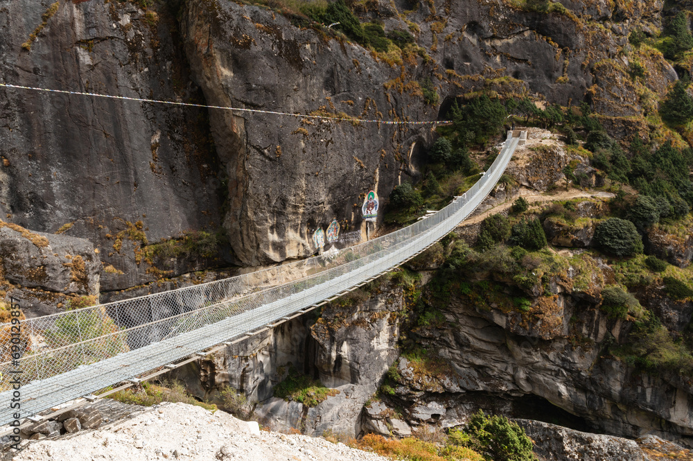 Fototapeta premium Landscape with Suspension Bridge over Bhote Kosi River during Three passes trekking in Sagarmatha national park, Nepal. A bridge with Guru Rinpoche images on rocks on the way to Thame.