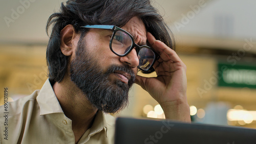 Indian bearded stressed man in glasses working at office with laptop Arabian businessman sad with business problem failure head pain suffering from chronic severe headache thinking trouble solution