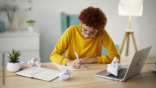 Young woman journalist writer in glasses writing article book on paper shits sitting at table near laptop. Crumpled papers on table around tired girl. Writing novel, letter, creating process concept. photo