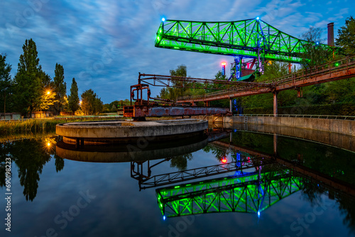 Old crane at a ruined factory complex called “Landschaftspark Nord“ in Duisburg-Meiderich – with reflection on the water surface of a clarifier at blue hour twilight. Colorful illumination at monument photo